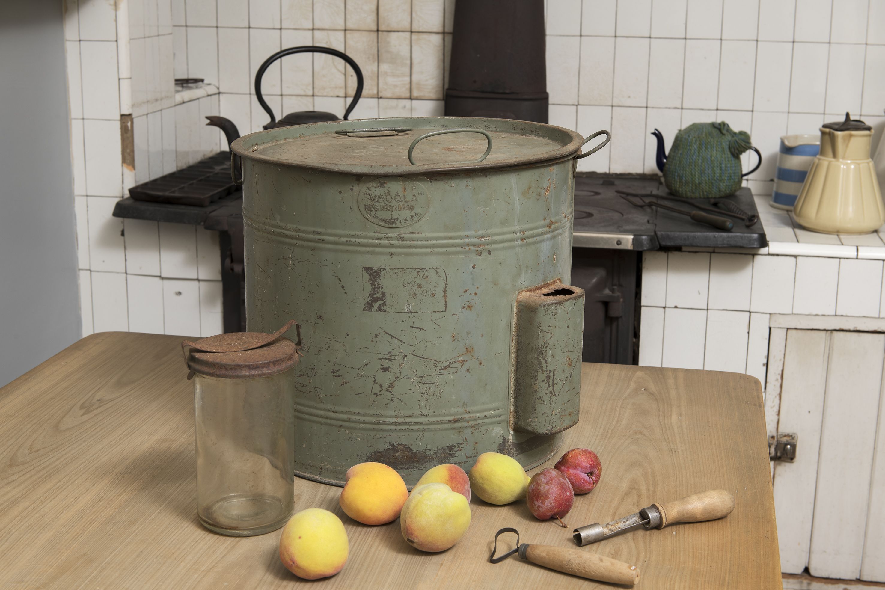 A green metal preserving pan sitting on a wooden table with fruit, a glass jar and utensils.