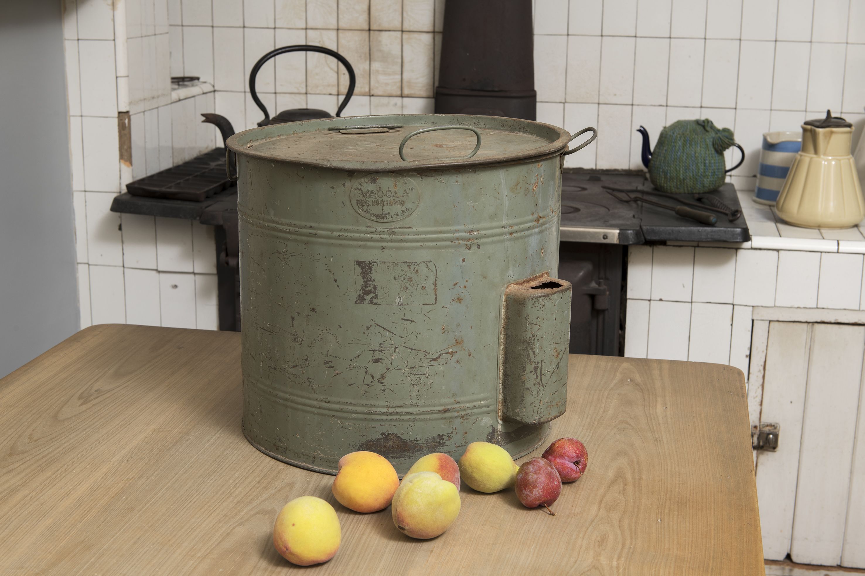A green metal preserving pan sitting on a wooden table with fruit in front.