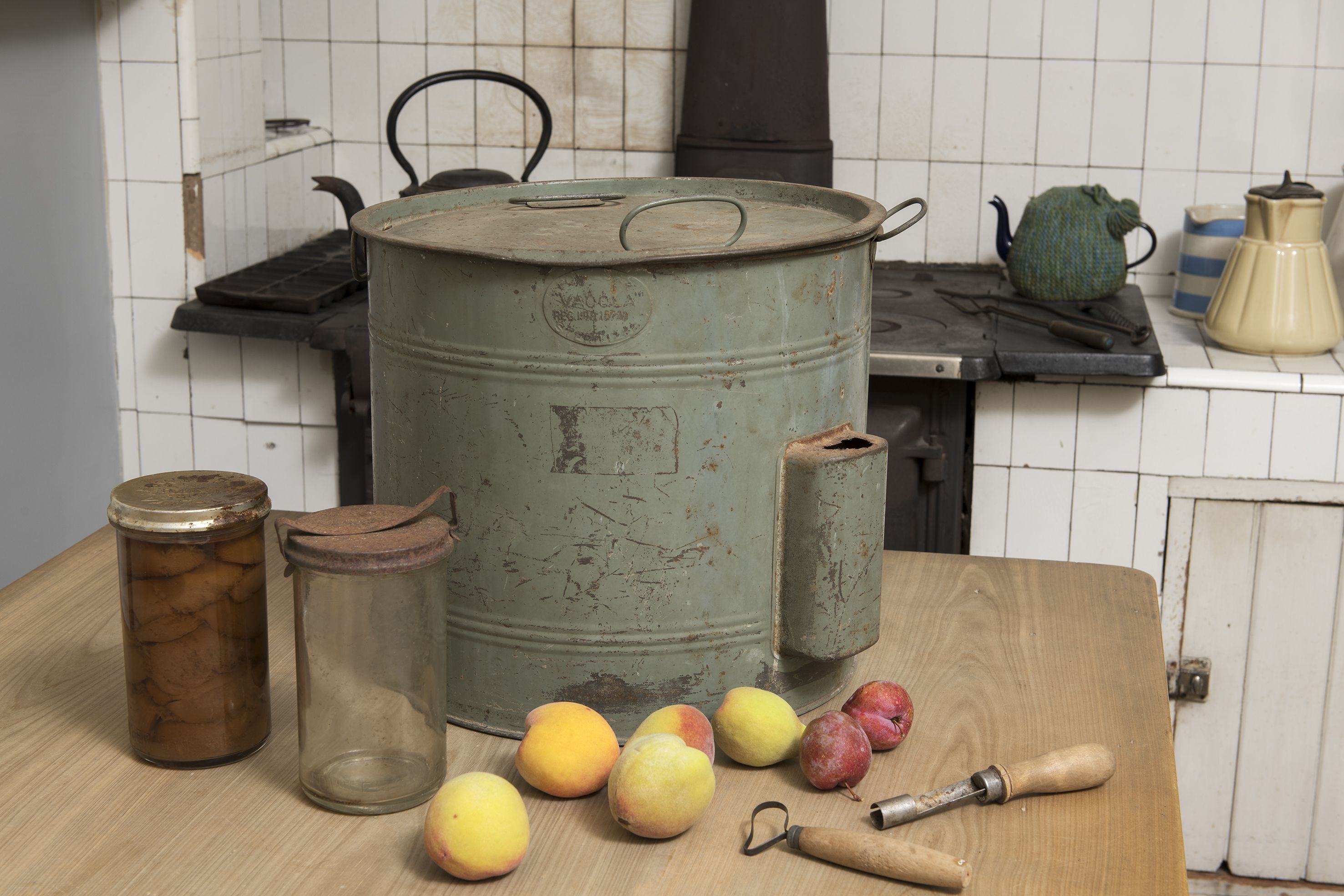 A green metal preserving pan sitting on a table, with fruit, glass jars, one with fruit, and utensils.